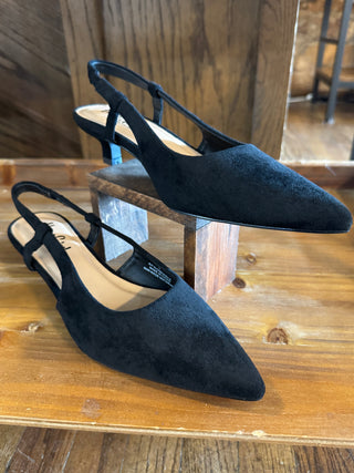 A pair of black suede slingback pointed-toe flats with low block heels, displayed on wooden blocks and a wooden table.