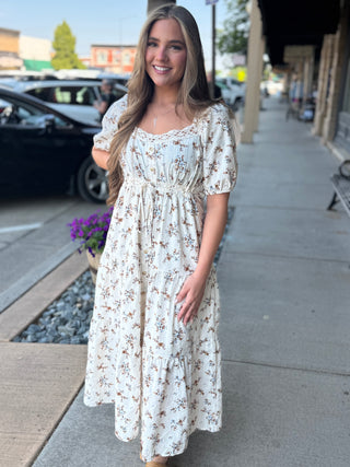 A woman stands on a city sidewalk wearing a cream-colored, short-sleeved midi dress with a brown and green floral print. The dress features a gathered bodice, a smocked waist, and a tiered skirt.