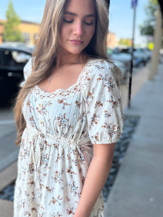 A woman wearing a short-sleeve cream-colored dress with a brown floral print, standing on a city sidewalk.