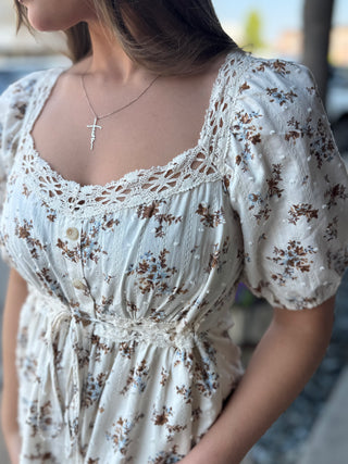 A close-up of a woman wearing a cream-colored short-sleeve dress with a brown and blue floral print, featuring lace trim along the scoop neckline and puff sleeves.