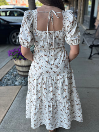 A woman viewed from behind wearing a cream-colored tiered midi dress with a brown and blue floral print, puff sleeves, and a tie-back neckline, standing on a sidewalk.