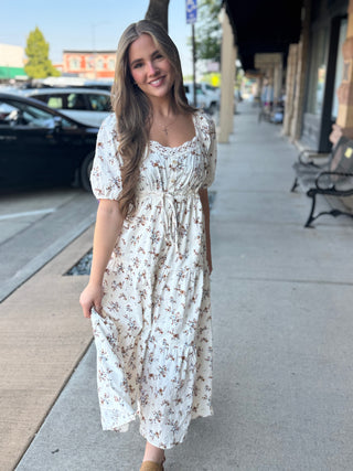 A woman wearing a short-sleeve cream maxi dress with a brown and pink floral print, standing on a sidewalk in front of a storefront.