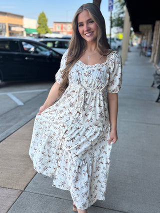 A woman wearing a white tiered midi dress with a brown floral print, standing on a city sidewalk.