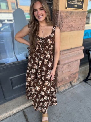 A woman wearing a brown sleeveless maxi dress with a beige and white floral pattern, standing outdoors next to a stone pillar.
