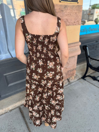 A woman viewed from behind wearing a brown sleeveless midi dress with a beige and white floral pattern, standing on a sidewalk in front of a storefront.