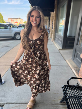A woman smiling and walking on a city sidewalk wearing a brown sleeveless maxi dress with a beige and white floral pattern.