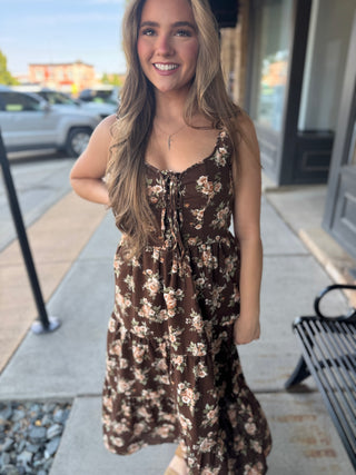 A woman wearing a brown floral midi dress with tie details at the neckline, standing on a sidewalk in front of a storefront.