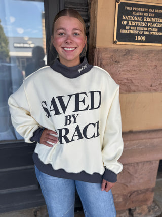 A smiling young woman wearing a cream-colored crewneck sweatshirt with dark gray ribbed trim and the text 'SAVED BY RACE' printed in black across the front, standing outdoors in front of a historic building wall.