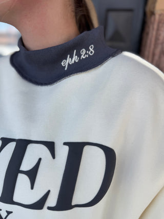 Close-up of a person wearing a cream sweatshirt with a navy mock neck collar featuring white embroidered text reading 'eph 2:8'.