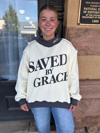 A smiling young woman stands outdoors wearing a cream-colored oversized sweatshirt with dark gray trim and the words 'SAVED BY GRACE' printed in large black letters across the front, paired with light blue jeans.