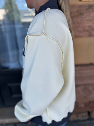 A person shown from the side wearing a cream-colored sweatshirt with dark navy ribbed collar, cuffs, and hem, photographed in an outdoor setting.