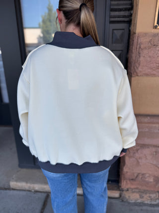 A person viewed from behind wearing a cream-colored oversized bomber-style sweatshirt with dark gray ribbed collar, hem, and cuffs, paired with blue jeans, standing outdoors in front of a building entrance.