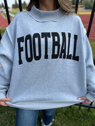 A person wearing an oversized light heather gray sweatshirt with the word 'FOOTBALL' printed across the chest in large black varsity-style letters, standing outdoors on grass near a track field.