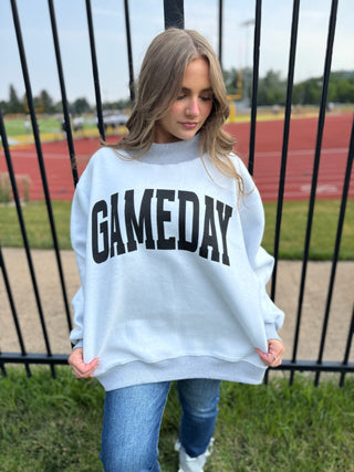 A woman wearing a light blue crewneck sweatshirt with 'GAMEDAY' printed in bold black letters across the chest, standing outdoors in front of a black metal fence at a sports field.