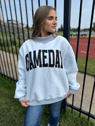 A woman wearing a light gray sweatshirt with 'GAMEDAY' printed in bold black letters across the chest, paired with blue jeans, standing outdoors near a black metal fence at what appears to be a track and field stadium.