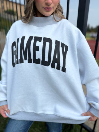 A woman wearing an oversized white sweatshirt with the word 'GAMEDAY' printed across the front in large black varsity-style letters, photographed outdoors near a fence.