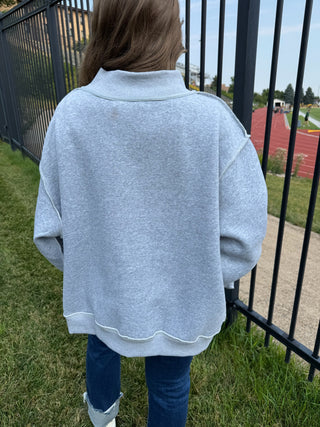 A person viewed from behind wearing an oversized light gray heather sweatshirt with a mock neck, paired with blue jeans, standing outdoors next to a black metal fence.