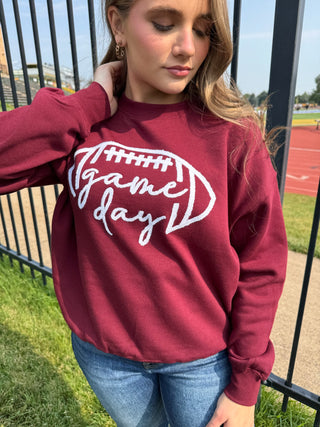 A woman wearing a maroon crewneck sweatshirt with a white graphic that reads 'Game Day' inside a football outline, paired with blue jeans, standing outdoors near a fence and track field.