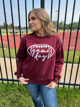 A woman wearing a maroon crewneck sweatshirt with a white graphic that reads 'game day' inside a football outline, standing in front of a black metal fence at an outdoor track and field.