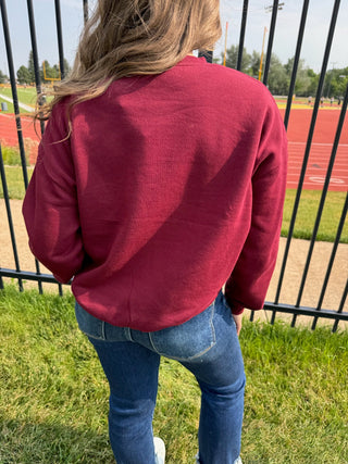 A person viewed from behind wearing a maroon crewneck sweatshirt and blue jeans, standing on grass in front of a black metal fence with a running track and field visible in the background.