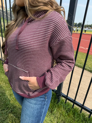 A woman wearing a burgundy and white striped hoodie with blue jeans, standing outdoors next to a black metal fence with a running track in the background.