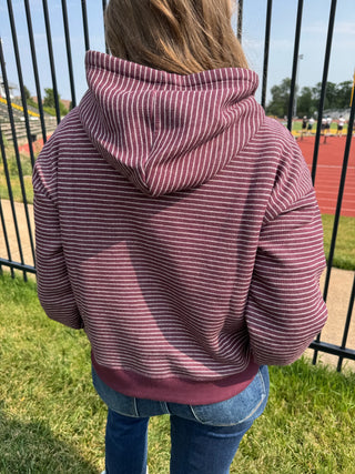 A person viewed from behind wearing a dusty rose and white horizontally striped hoodie, standing outdoors on a grassy field with a black metal fence and a running track in the background.