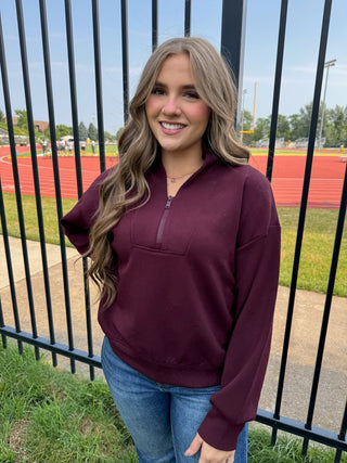 A woman with long wavy blonde hair stands outdoors in front of a black metal fence, wearing a maroon long-sleeve quarter-zip pullover sweatshirt and blue jeans. The background shows a school track field under a blue sky.