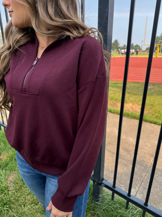 A woman wearing a long-sleeve half-zip sweatshirt in deep burgundy, paired with blue jeans, standing outdoors next to a black metal fence with a track and field visible in the background.