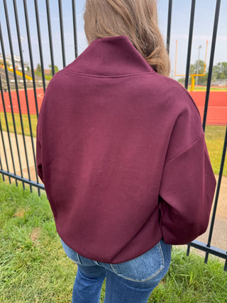 A person viewed from behind wearing a maroon crewneck sweatshirt and blue jeans, standing outdoors in front of a black metal fence with a running track and field visible in the background.