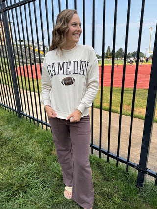 A woman stands outdoors on a grassy field behind a black metal fence, wearing a cream sweatshirt with 'GAME DAY' and a football graphic, paired with mauve wide-leg pants and white slide sandals.