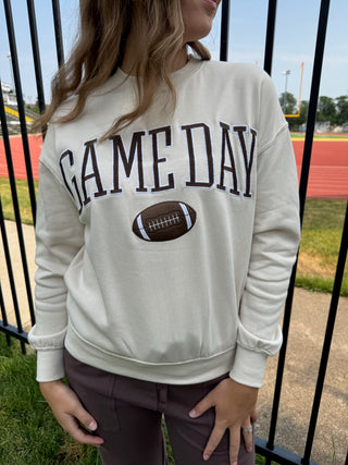 A woman wearing a cream-colored crewneck sweatshirt with 'GAME DAY' and a football graphic printed on the front, standing outdoors in front of a black fence at a sports field.
