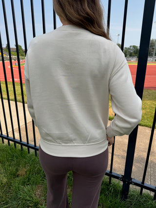 A person viewed from behind wearing a cream-colored crewneck sweatshirt and mauve-brown leggings, standing outdoors in front of a black metal fence with a running track and field visible in the background.
