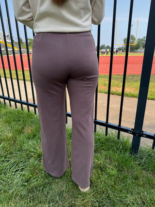 A person viewed from behind wearing dusty mauve wide-leg sweatpants and a cream sweatshirt, standing on grass in front of a black metal fence with a running track and field visible in the background.