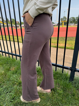 A person wearing dusty mauve wide-leg sweatpants, standing outdoors on grass in front of a black metal fence with a running track in the background.