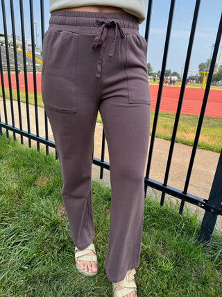 A person wearing loose, straight-leg drawstring sweatpants in a muted mauve-brown color, standing outdoors on grass near a black metal fence and a running track.
