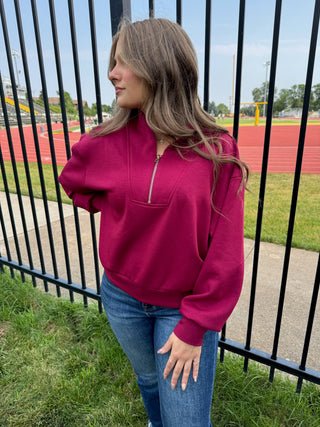 A woman wearing a magenta half-zip sweatshirt and blue jeans stands outdoors next to a black metal fence with a running track and field visible in the background.
