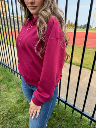 A woman wearing a bright magenta half-zip sweatshirt and blue jeans, standing outdoors next to a black metal fence with a track and field stadium in the background.
