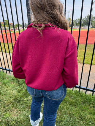A person viewed from behind wearing a bright magenta-pink cropped hoodie and blue jeans, standing outdoors on grass in front of a black metal fence with a running track field in the background.