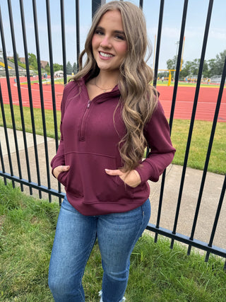 A woman with long wavy blonde hair stands outdoors in front of a black metal fence, wearing a burgundy half-zip pullover hoodie with a kangaroo pocket and blue jeans.