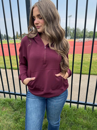 A woman wearing a burgundy quarter-zip pullover sweatshirt with front pockets and blue jeans, standing outdoors in front of a black metal fence with a running track and field visible in the background.