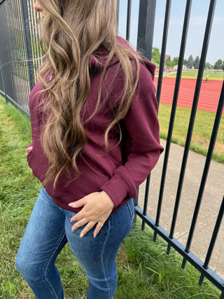A woman wearing a burgundy pullover hoodie and blue jeans, standing outdoors next to a black metal fence with a running track in the background.