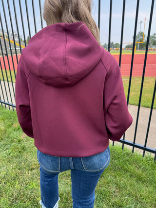 A person viewed from behind wearing a cropped maroon hoodie and blue jeans, standing outdoors on grass near a black metal fence with a track and field visible in the background.