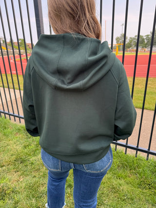 A person viewed from behind wearing an oversized dark green hooded sweatshirt and blue jeans, standing outdoors on grass in front of a black metal fence with a running track in the background.