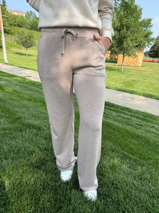 A person standing on a grassy lawn wearing light beige heather sweatpants with a drawstring waist and a matching sweatshirt, with trees and a walkway in the background.