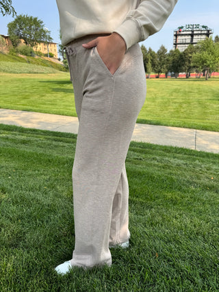 A person wearing light beige heather sweatpants and a matching sweatshirt stands outdoors on a green grassy lawn with a sidewalk and trees in the background.