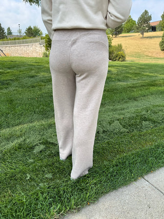 A person standing outdoors on a grassy lawn wearing light beige heather sweatpants and a matching sweatshirt, shown from the back.