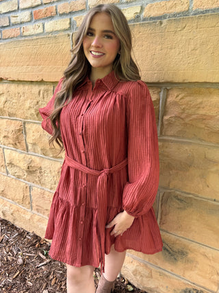 A woman wearing a rust-colored, long-sleeve crinkle-textured mini dress with a button-up front and matching tie belt, standing against a tan brick wall.