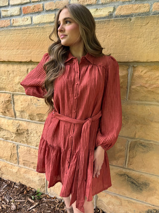 A woman wearing a rust-red pleated long-sleeve shirt dress with a matching tie belt, standing against a tan brick wall.