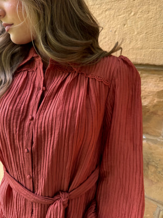 A close-up of a woman wearing a rust-red crinkle-textured long-sleeve shirt dress with a tie waist and lace detail at the collar, set against a textured beige wall.