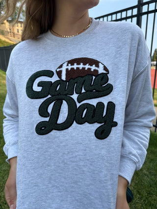 A person wearing a light heather gray long-sleeve sweatshirt with a large 'Game Day' graphic on the front, featuring a brown and white football above dark green script lettering, photographed outdoors on grass.